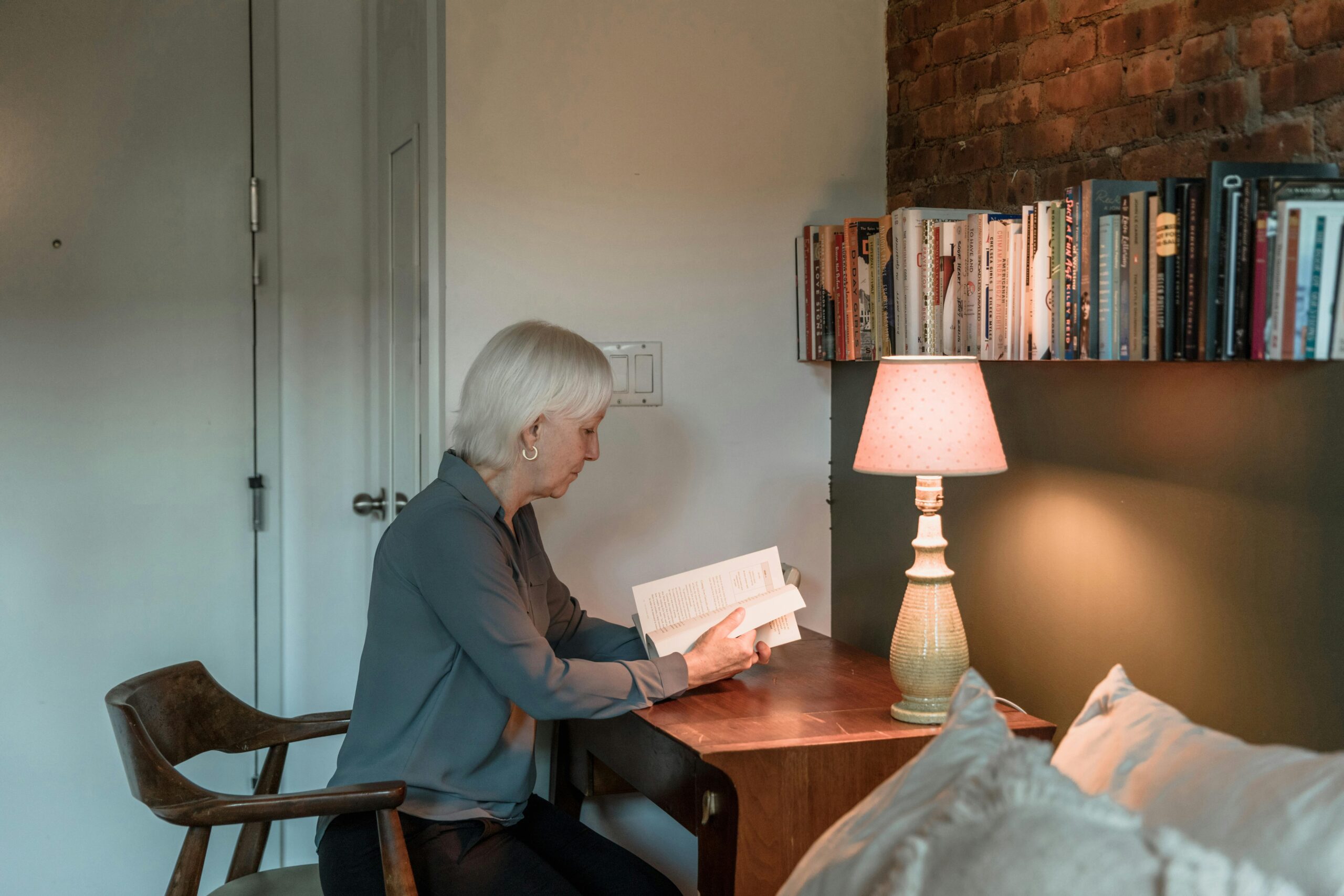 Elderly woman reading a book by a lamp in a cozy home setting.